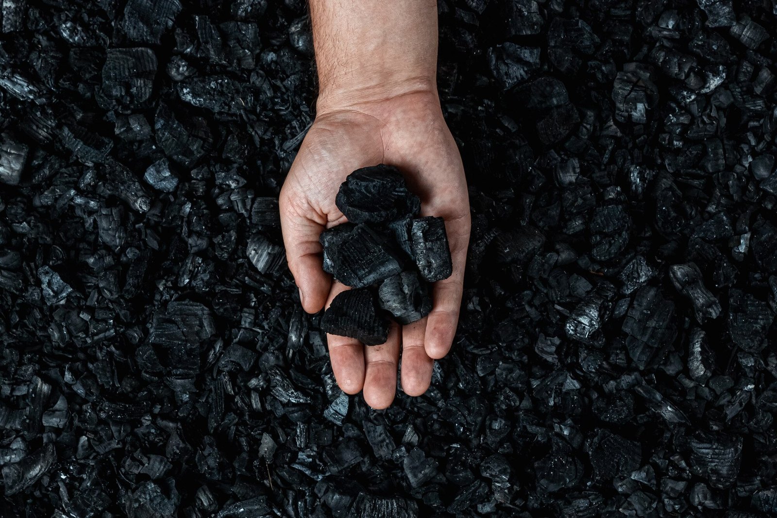 Hand holding biochar chunks over a bed of charcoal pieces. The biochar appears as dark, porous black material representing the carbon-rich product used for soil enhancement and carbon sequestration. The image conveys the tangible, industrial nature of the biomass product against the darker charcoal background.